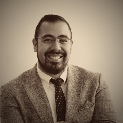 Portrait of Amirali Majidi in a grey tweed blazer and tie, smiling